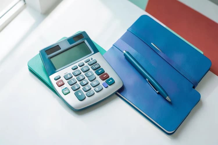 Calculadora com números na tela e uma mão anotando em um caderno.