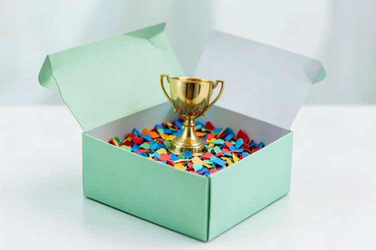 Caixa de presente aberta com confetes coloridos e um pequeno troféu dourado saindo dela.